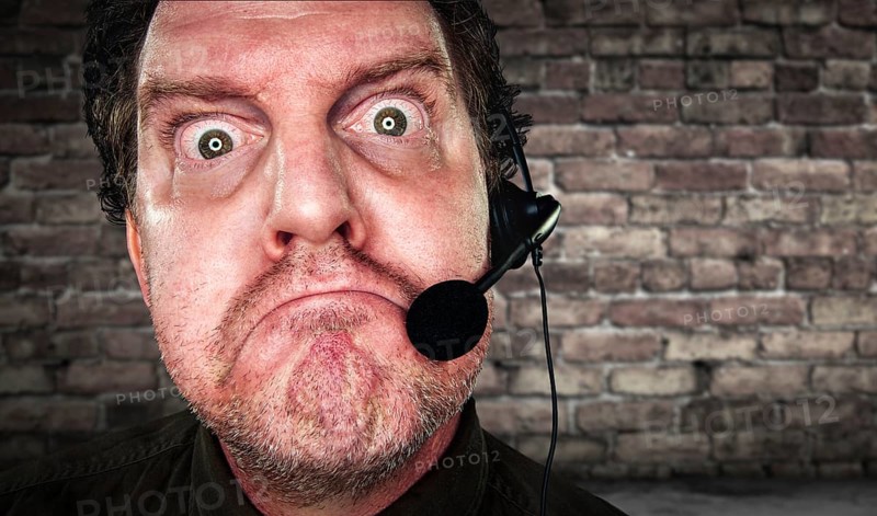 Grumpy businessman with frowning face wearing phone headset in office with brick background