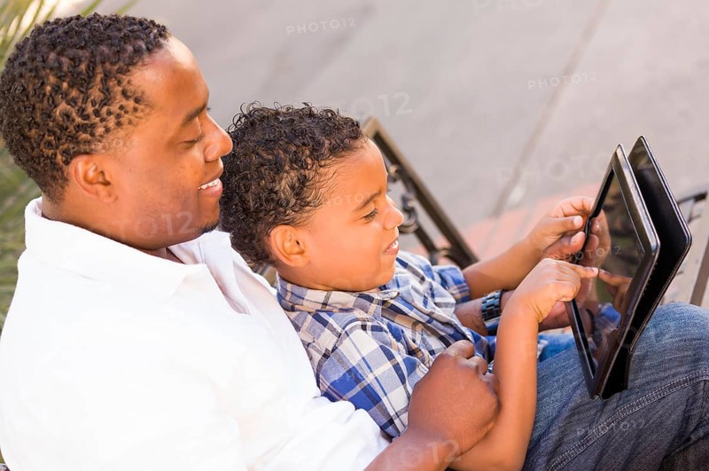 African american father and mixed-race son using computer tablet on bench in park