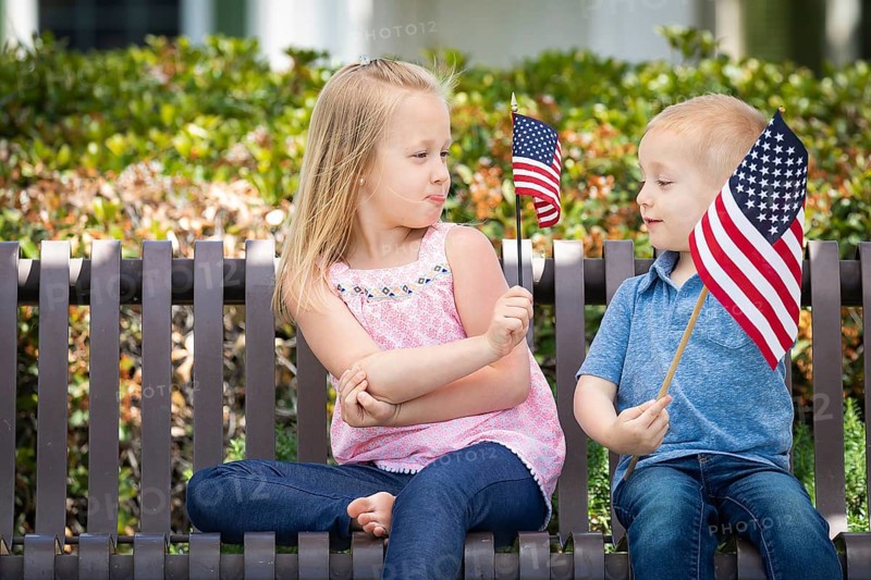 Young sister and brother comparing each others american flag size on the bench at the park