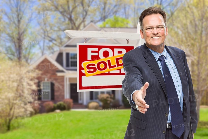 Smiling male agent reaching for a hand shake in front of beautiful house and sold real estate sign