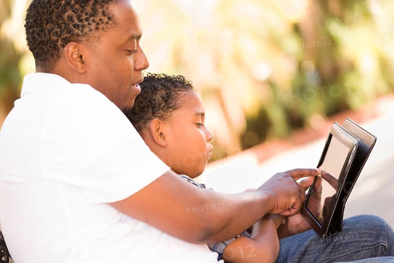 African american father and mixed-race son using computer tablet on bench in park