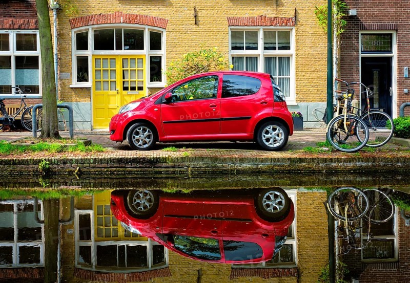 Red car on canal embankment in street of Delft with reflection and bicycle
