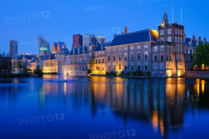 View of the Binnenhof House of Parliament and the Hofvijver lake with downtown skyscrapers in background illuminated in the evening. The Hague