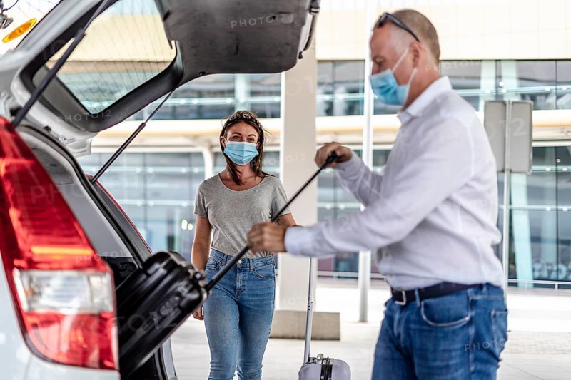 A taxi or Uber driver helping a passenger in a protective mask with her luggage at the airport