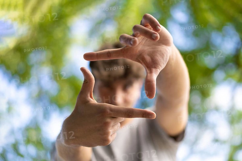 A handsome young man blinks his eyes and looks through a frame made up of hands. Attractive man make frame by fingers