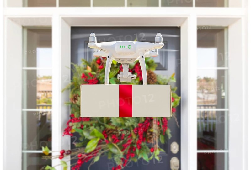 Drone delivering wrapped package with red ribbon to christmas decorated house porch