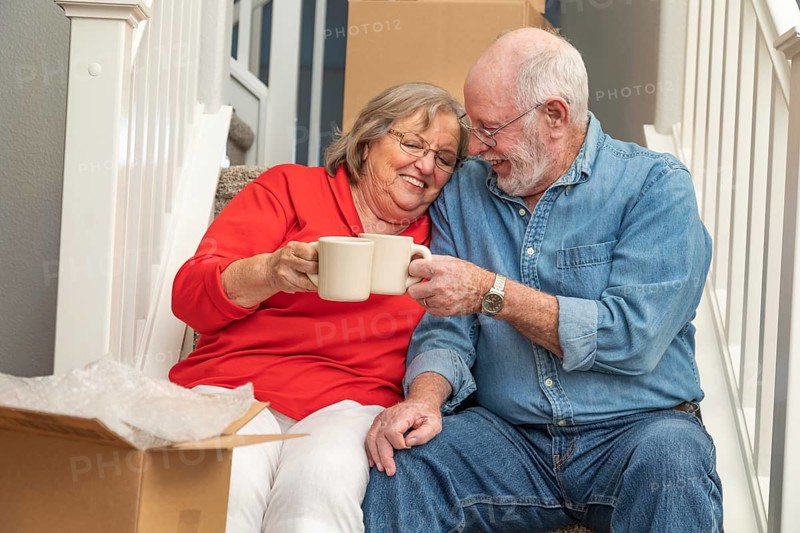 Tired senior adult couple resting on stairs with cups of coffee surrounded with moving boxes