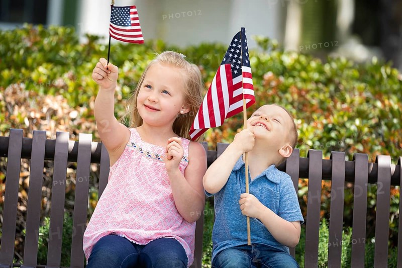 Young sister and brother comparing each others american flag size on the bench at the park