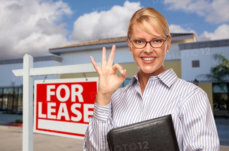 Smiling businesswoman with okay sign in front of vacant office building and for lease real estate sign