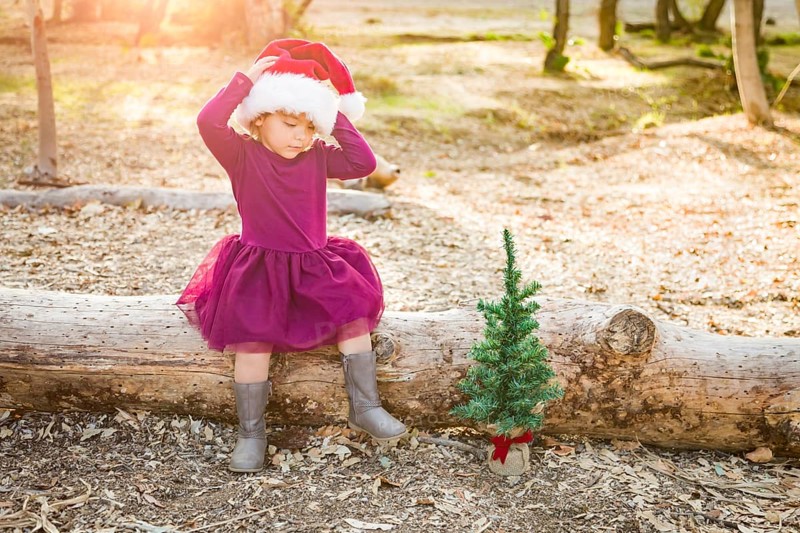 Cute mixed-race young baby girl having fun with santa hat and christmas tree outdoors on log