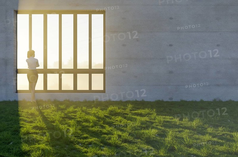 Woman standing at window with bright sun light shining through concrete wall in grass and rocks field