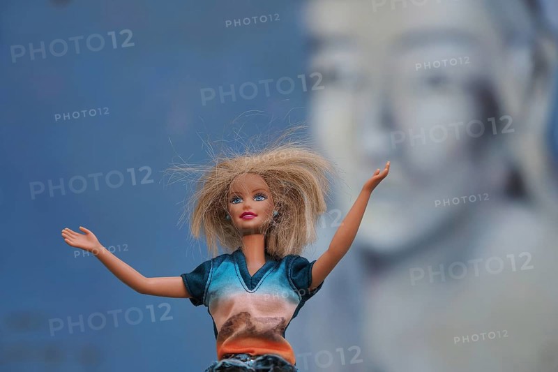 Barbie doll with the gesture of outstretched arms in front of wall with Buddha head