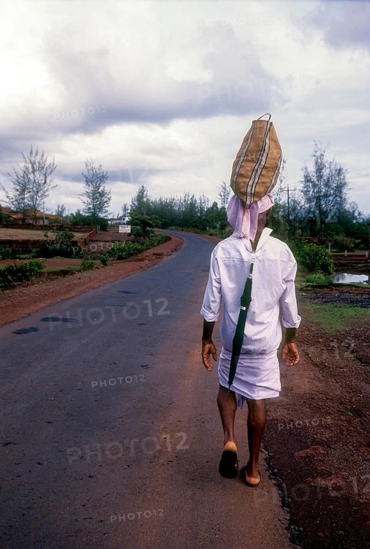 A man carrying bag over his head and an umbrella behind his shirt on the road