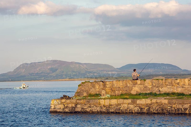 Man rod fishing on summers evening as boat heads out to sea. Sligo