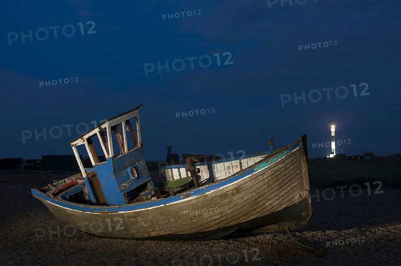 Abandoned fishing boat on shingle beach with distant lighthouse at night
