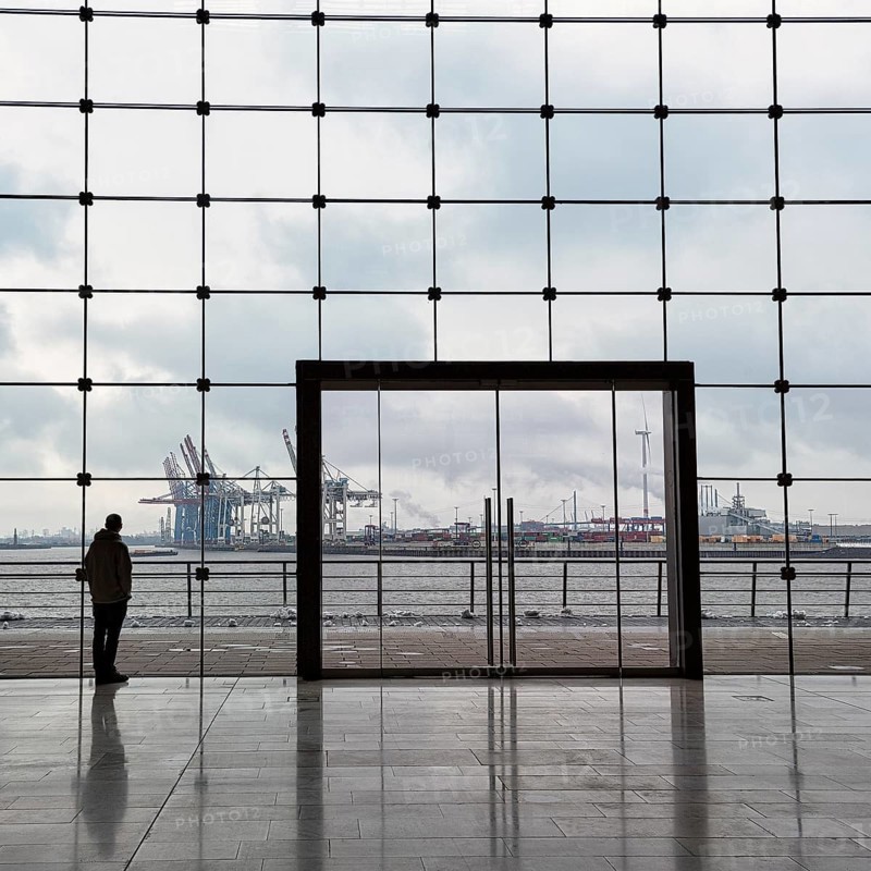 Young man standing in front of modern glass facade with large door and looking at the harbour