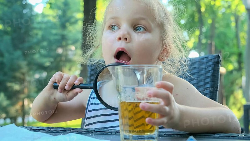 Little girl looks into the lens around. Close-up of blonde girl studying the world around her looking at it through magnifying glass while sitting in street cafe on the park