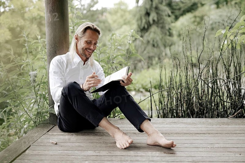 Businessman in white shirt sits on wooden jetty and writes in a book,