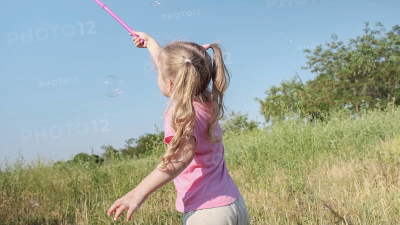 Little girl catches soap bubbles in butterfly net while playing in tall grass in park. Cute little girl smiles and catches on soap bubbles in aerial insect net in meadow on sun day. Odessa, Ukraine