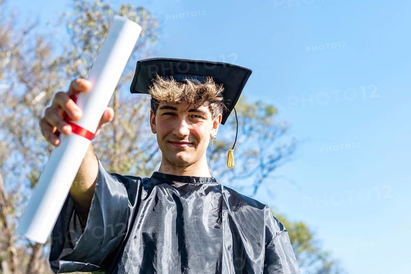 Happy graduated young man wearing a bachelor gown and a black mortarboard and showing his diploma,