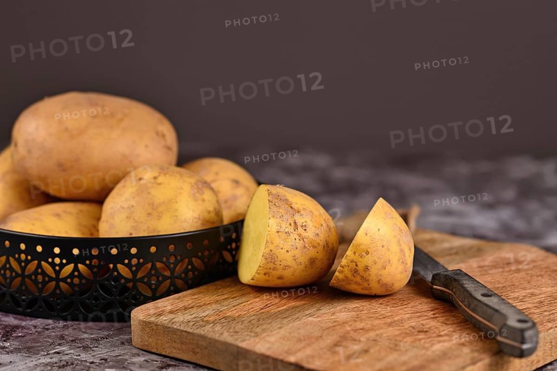 Cutting raw potato with peel on wooden cutting board with pile of whole potatoes in background,