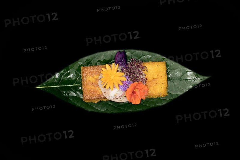Fried polenta slice with a slice of lotus root and meadow flowers as decoration on an avocado leaf, foddfotography with black background