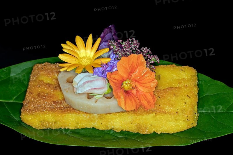Fried polenta slice with a slice of lotus root and meadow flowers as decoration on an avocado leaf, foddfotography with black background