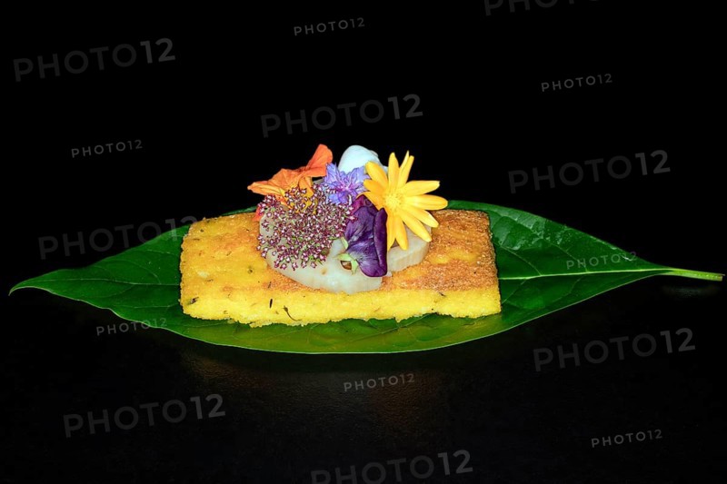 Fried polenta slice with a slice of lotus root and meadow flowers as decoration on an avocado leaf, foddfotography with black background