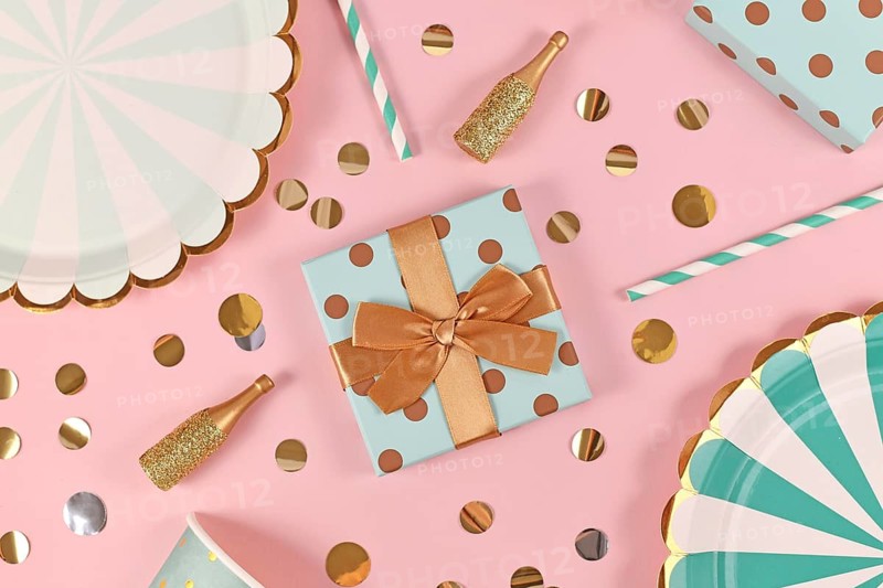 Birthday gift box with with teal blue paper plates, small golden champagne bottles and confetti on pink background