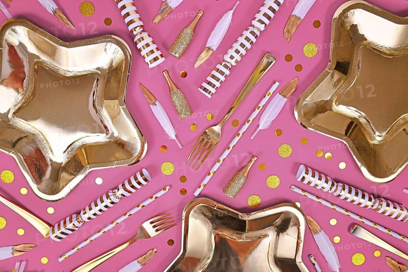 Party flat lay with golden and white decoration like plates, confetti and feathers on pink background