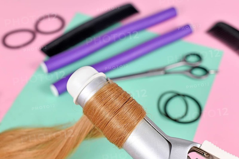 Strand of blond hair wrapped around curling iron with hair styling tools in blurry background,