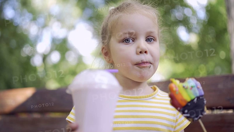 Ittle girl eats a colorful gingerbread and holds a milkshake in her hand. Close-up of cute child girl sitting on park bench and eating cookies with a milkshake and looks into the camera lens. Odessa, Ukraine