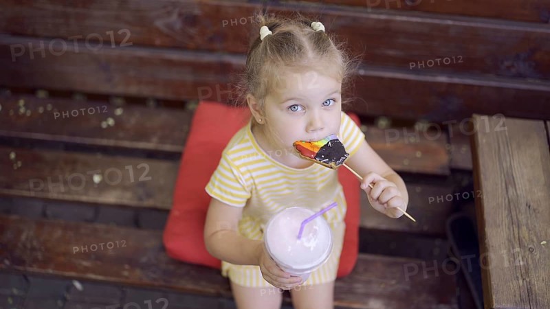Ittle girl eats a colorful gingerbread and holds a milkshake in her hand. Close-up of cute child girl sitting on park bench and eating cookies with a milkshake and looks into the camera lens. Top view. Odessa, Ukraine