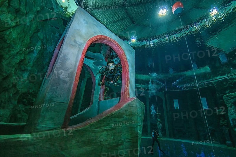 Female diver dives in 10 metres depth in indoor diving tower through imitated bridge house over bow of stylised small ship