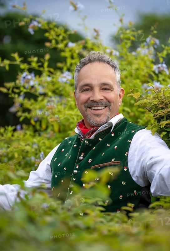 Laughing middle-aged man in Bavarian traditional costume standing by a hedge