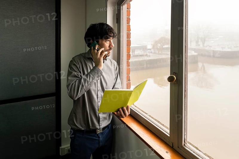 Latino businessman working in an office overlooking the river. Talking on the phone and reviewing notes while he looks out the window