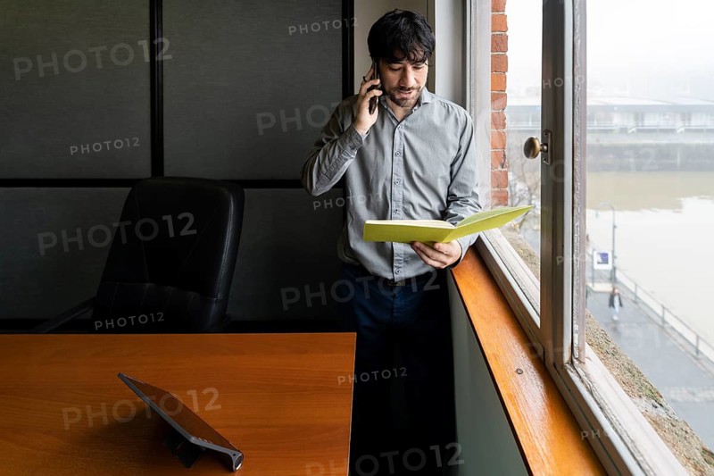 Latino businessman working in an office overlooking the river. Talking on the phone and reviewing notes