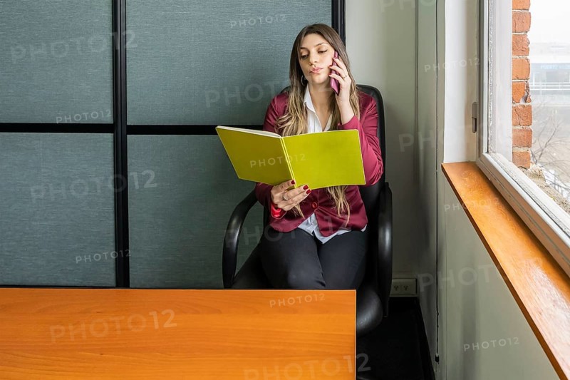 Young blonde business woman sitting in her office using the phone while checking notes