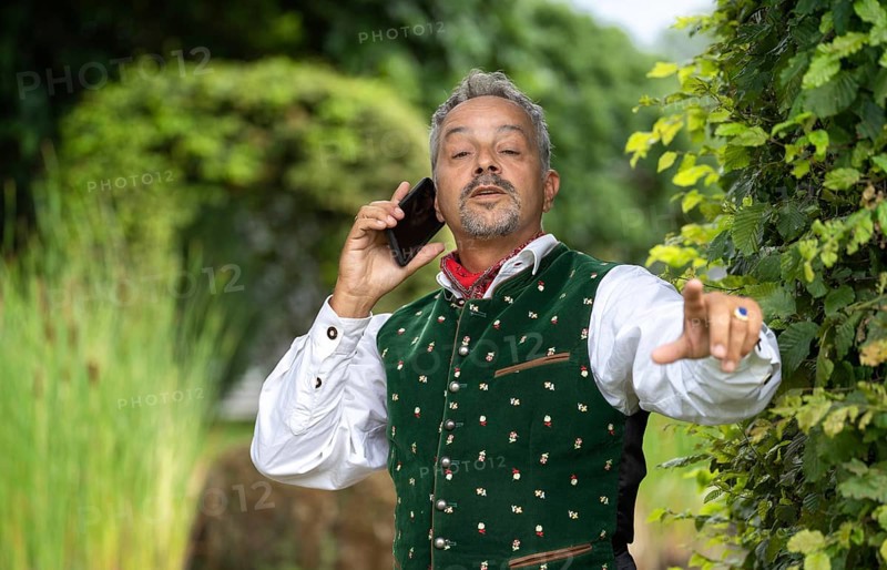 Man with telephone in Bavarian traditional costume at a hedge