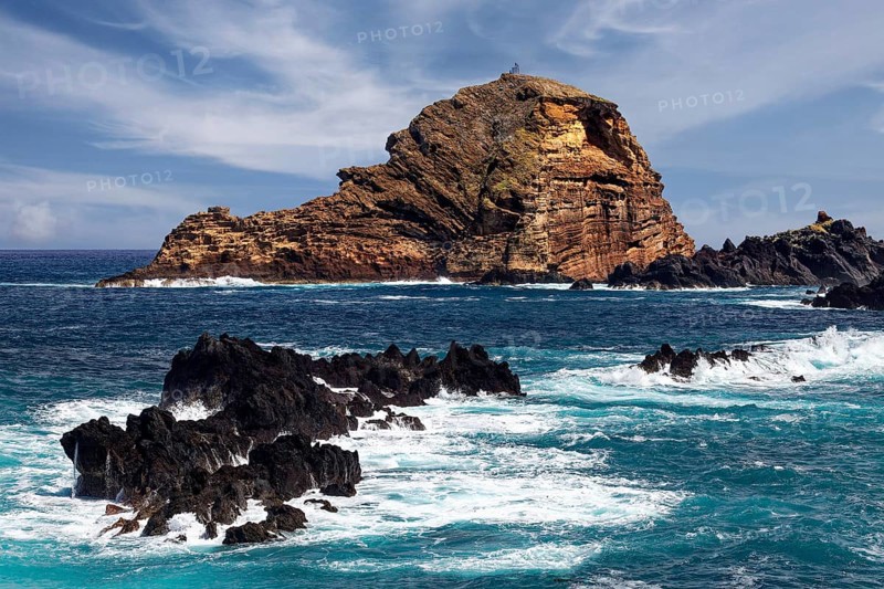 Rocks unswept by surf at the back small rocky island Ilheu Mole with lighthouse Farol do Porto Moniz