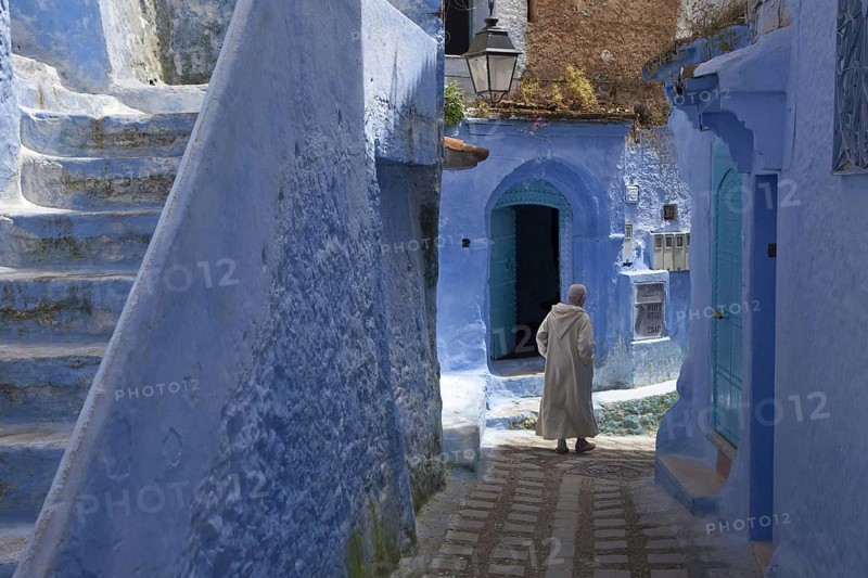 Man with djellaba walking next to blue buildings and staircase in an alley of the city