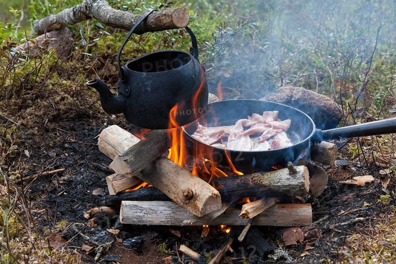 Blackened tin kettle boiling water and pan cooking bacon over flames from campfire