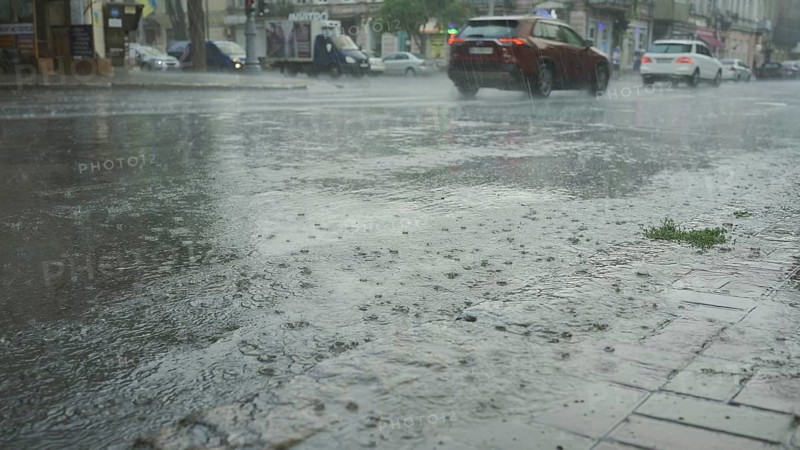 Raindrops fall on the road with cars driving in the background. Heavy torrential rain in the city