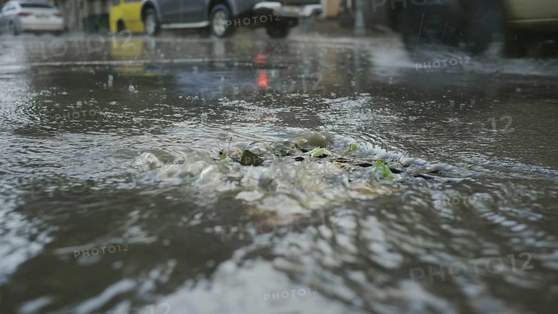 A stream of rainwater flows into the storm drain cars are driving in the background. Heavy torrential rain in the city