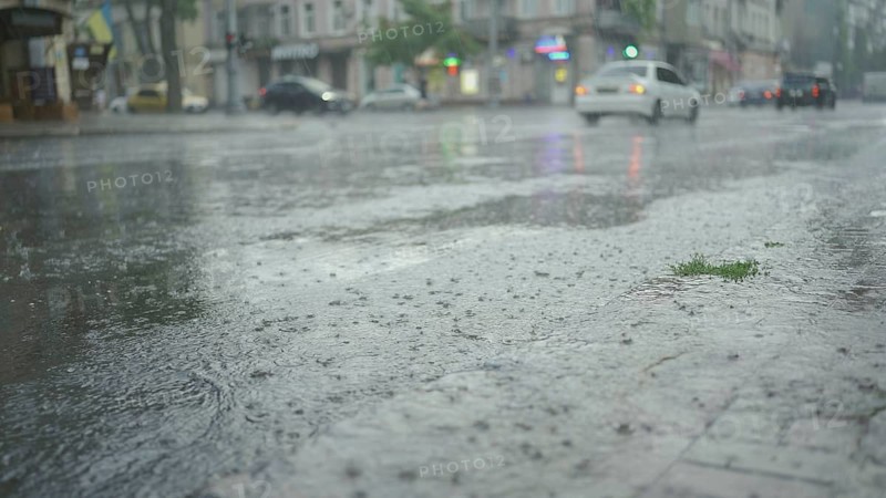 Raindrops fall on the road with cars driving in the background. Heavy torrential rain in the city