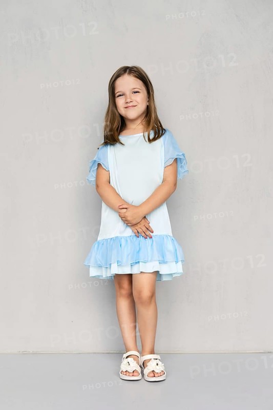 Cute little girl in blue dress and white sandals posing with hands clasped down near the gray wall