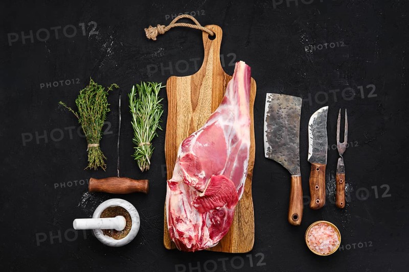 Fresh raw lamb leg bone in on wooden cutting board with herbs and seasoning