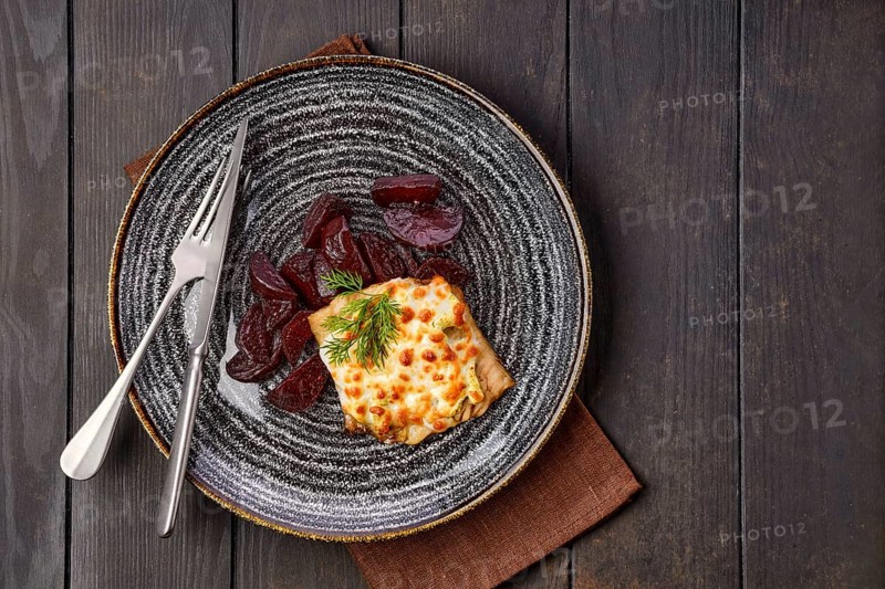 Top view of fried cod fillet with melted cheese topping and roasted beetroot slices on dark wooden table