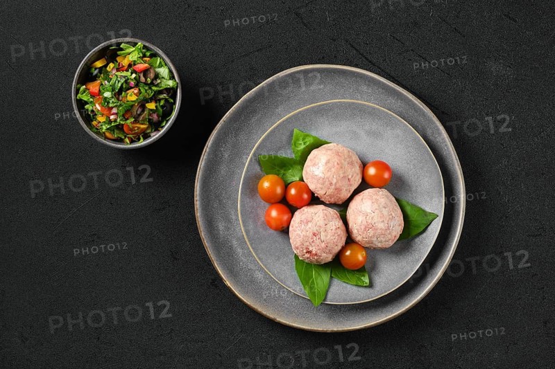 Top view of semifinished frozen veal meatballs on a plate with basil and tomato