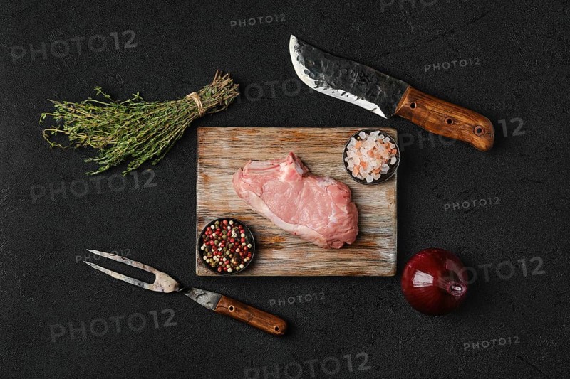 Overhead view of raw veal steak bone in with ingredients for cooking on black background